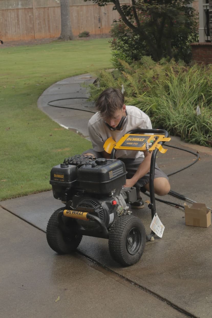 Man inspecting a Dewalt power washer on a driveway, surrounded by a well-maintained lawn.