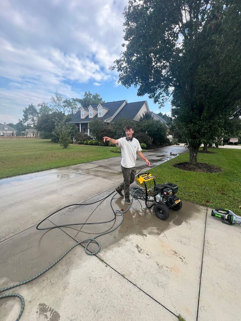 Man using a power washer to clean a driveway with a house and trees in the background.